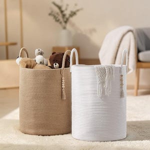 May include: Two cylindrical rope baskets, one in tan and one in white, with arched handles. The tan basket contains soft toys. The white basket holds a white blanket. Both baskets have beaded tassels.