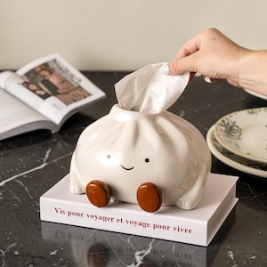 May include: A novelty tissue box in the shape of a dumpling, made from off-white ceramic. It features a smiling face and brown feet. The box rests on a white base with the text "Vis pour voyager et voyage pour vivre". A hand is pulling a tissue.