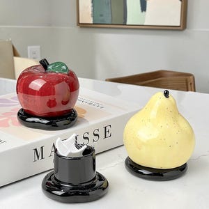 Puede incluir: Tres recipientes de cerámica con forma de fruta: una manzana roja, una pera amarilla y un recipiente cilíndrico negro. La manzana y la pera se asientan sobre bases negras. Los objetos están sobre una superficie blanca.