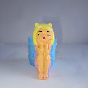 Puede incluir: Una figura caprichosa de una mujer con cabello amarillo y ojos cerrados, apoyando la barbilla en sus manos. Lleva un vestido rosa y está frente a un barril azul. La figura está pintada con colores brillantes.