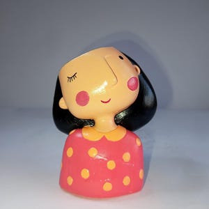 Może przedstawiać: Ceramiczna figurka dziewczynki z czarnymi włosami, różowymi policzkami i uśmiechniętą miną. Ma na sobie czerwoną sukienkę w żółte grochy i żółty kołnierzyk. Figurka jest przedmiotem dekoracyjnym.