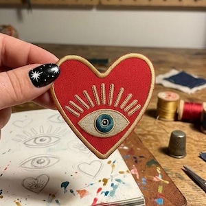 May include: A red heart-shaped embroidered patch with a gold border. The patch features an eye design with a blue iris and radiating lines. The patch is held in a hand with black nail polish with white star designs. The background includes sewing supplies.