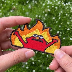 May include: Embroidered patch featuring a red character engulfed in flames with arms raised. The patch has a yellow and orange flame design, with a black outline. The character has white eyes and a black mouth.