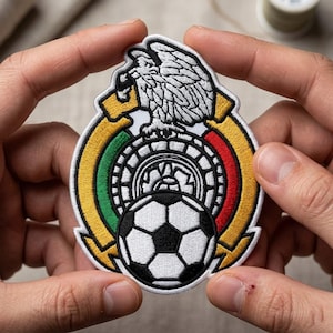 May include: A circular embroidered patch featuring a white eagle perched on a cactus, positioned above a black and white soccer ball. The design includes a gold, green, and red border with intricate stitching details throughout the emblem.