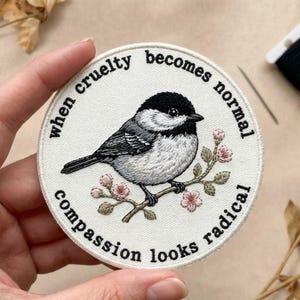 May include: Embroidered patch featuring a detailed bird perched on a flowering branch. The patch is circular with the text "when cruelty becomes normal, compassion looks radical" in a black font. The bird is black, white, and gray.