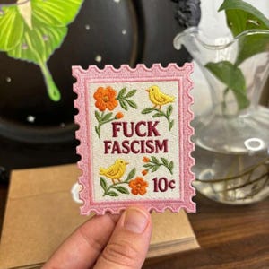 May include: Embroidered patch in a postage stamp design with a pink border. The patch features the text "FUCK FASCISM", yellow birds, and orange flowers. The patch also includes the number "10c".