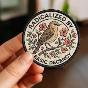 May include: Round embroidered patch with a bird perched on a branch with flowers. The patch has the text "RADICALIZED BY BASIC DECENCY" in a circular design. The bird is brown and beige, and the flowers are pink.