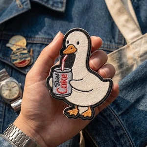 May include: Embroidered patch featuring a white cartoon duck holding a Diet Coke can with a red straw. The duck has an orange beak and feet, outlined in black. The text "Diet Coke" is visible on the can. The patch is on a denim background.