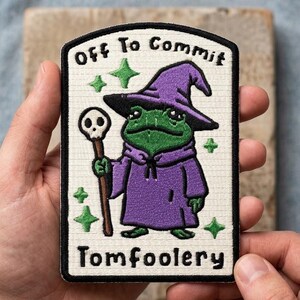 May include: A rectangular patch featuring a green frog wizard in a purple robe and hat, holding a staff. The patch has the text "Off To Commit" above the frog and "Tomfoolery" below. Green star shapes are scattered around the frog.