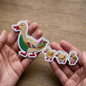 May include: Embroidered duck patch set featuring a mother duck with three ducklings. The patches have a white border, with the mother duck in tan, green, and pink, and the ducklings in yellow and brown. The ducks have red beaks and feet.