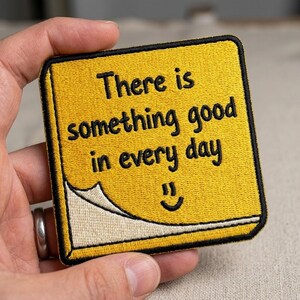 May include: A square yellow embroidered patch with a black border, featuring the black text "There is something good in every day" and a small black smiley face. The bottom left corner shows a white paper-like peel effect.