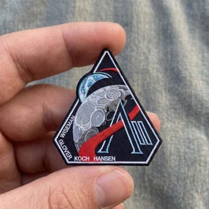 May include: A black, triangular patch featuring a stylized moon, Earth, and red and white accents. The patch includes the names "WISEMAN", "GLOVER", "KOCH", and "HANSEN". The design is reminiscent of a space mission emblem.