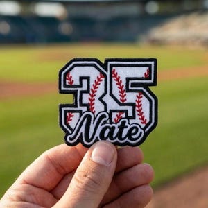 May include: A baseball-themed patch with the number "35" in white, outlined in black, and featuring red stitching to mimic a baseball. The name "Nate" is embroidered in a script font below the number. The patch is held in front of a blurred baseball field.