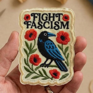 May include: Embroidered patch with the words "FIGHT FASCISM" in black lettering. A blue bird is perched on a branch with red flowers. The patch has a cream-colored border and is held in a hand.