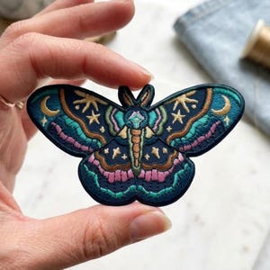 May include: Embroidered moth patch in shades of teal, navy, pink, and gold. The patch features a detailed design of a moth with celestial elements like stars and a crescent moon. The patch is held between fingers.