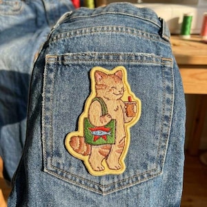May include: Light blue denim jeans with a decorative patch. The patch features a cartoon cat holding a drink and a bag with a fish design. The patch is outlined in yellow.