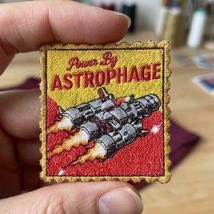 May include: Embroidered patch with a yellow and red background. The patch features a spaceship design with the text "Power By ASTROPHAGE". The spaceship is gray with orange flames.