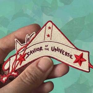 May include: Embroidered patch with the text "Savior of the Universe" in black lettering on a cream-colored banner. The patch features red star accents and a red decorative design. The patch is held in a hand.
