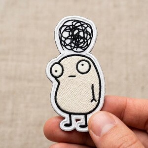 May include: Embroidered patch featuring a cartoon character with a beige body, large eyes, and a scribbled black design above its head. The patch has a white border and is held by a hand.