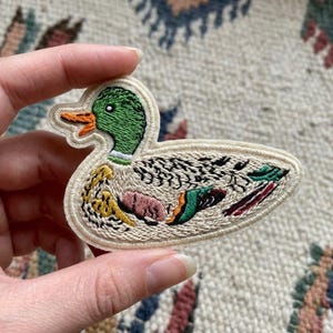 May include: Embroidered duck patch with a green head, orange beak, and cream-colored border. The duck's body features detailed stitching in various colors, including yellow, pink, and green. This patch is ideal for adding a decorative touch to clothing or accessories.