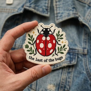 May include: An embroidered patch featuring a red ladybug with white spots on a cream background, surrounded by green leaves and small gold stars. The text "the last of the bugs" is stitched in black at the bottom of the scalloped-edge patch.