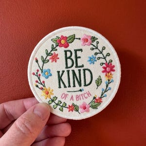 May include: Round embroidered patch with the text "BE KIND of a BITCH" in dark green. The patch is cream-colored with a floral wreath in pink, yellow, blue, and red. The patch is held by a hand against a red background.
