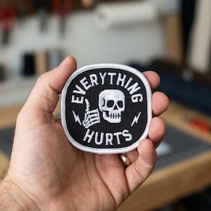May include: A black and white embroidered patch with the words "EVERYTHING HURTS" surrounding a skull and crossbones design. The patch has a white border and a lightning bolt on each side.