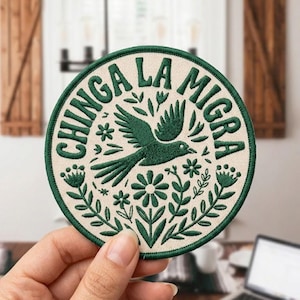 May include: Round embroidered patch with a cream background and a green border. The patch features the text "CHINGA LA MIGRA" in green, along with a green bird and floral design. The patch is held by a hand.