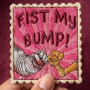 May include: Embroidered patch with the text "FIST MY BUMP!" in maroon on a pink background. The patch features two fists bumping, one white and one tan, with a cream-colored border.