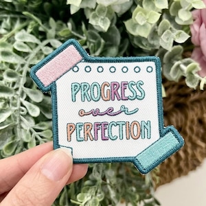 May include: A square embroidered patch shaped like a notepad with a teal border. The white center features the text PROGRESS over PERFECTION in teal, pink, and orange thread. A pink tab is at the top left and a teal tab is at the bottom right.