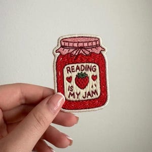 May include: Embroidered patch in the shape of a jam jar. The jar is red with a pink lid and a white label that reads "Reading is my jam" with a strawberry and hearts. The patch has a cream-colored border.