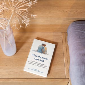 May include: A book titled "When the Game Gets Soft" rests on a wooden surface, beside a vase with dried flowers. The cover shows an illustration of two people embracing. The book's subtitle is visible.