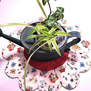 May include: A green watering can with a white and red flower-shaped pot holder. The pot holder has a white background with a red border and a variety of colorful floral and animal designs. The watering can is filled with a green plant.