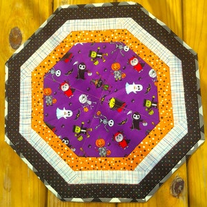 May include: A purple and orange octagon-shaped table runner with a Halloween theme. The runner features a variety of Halloween characters, including ghosts, witches, bats, and pumpkins. The runner is made of fabric and has a quilted design.