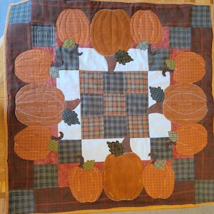 Puede incluir: Un camino de mesa acolchado con un motivo de calabaza. El camino de mesa tiene forma cuadrada con calabazas naranjas, hojas verdes y tallos marrones. El fondo es un patchwork de telas a cuadros y lisas en tonos marrones, naranjas y verdes.