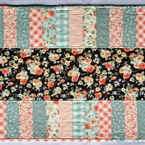 May include: A quilted table runner with a patchwork design featuring floral, gingham, and striped patterns in shades of pink, blue, and white. The center panel is a black fabric with a floral print.