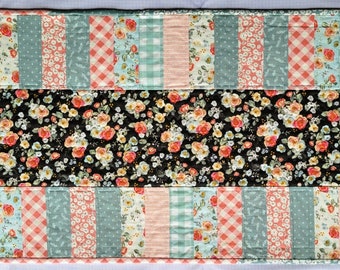 May include: A quilted table runner with a patchwork design featuring floral, gingham, and striped patterns in shades of pink, blue, and white. The center panel is a black fabric with a floral print.