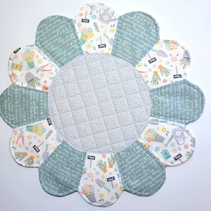 May include: A gray and white quilted table runner with a flower-shaped design. The runner is made of fabric with a floral print and text that says "Sunshine Home" and "Flowers".