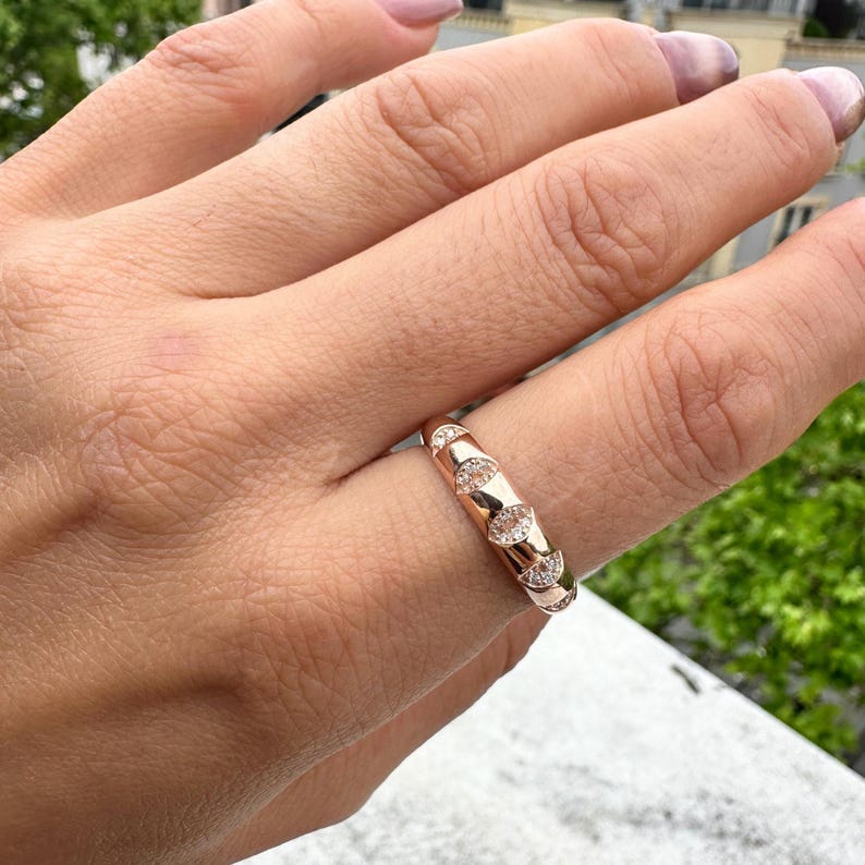 Puede incluir: Un anillo de oro rosa con un patr&oacute;n repetitivo de &oacute;valos, cada uno lleno de peque&ntilde;as piedras preciosas transparentes. El anillo est&aacute; en un dedo, con un fondo borroso. Es una joya.