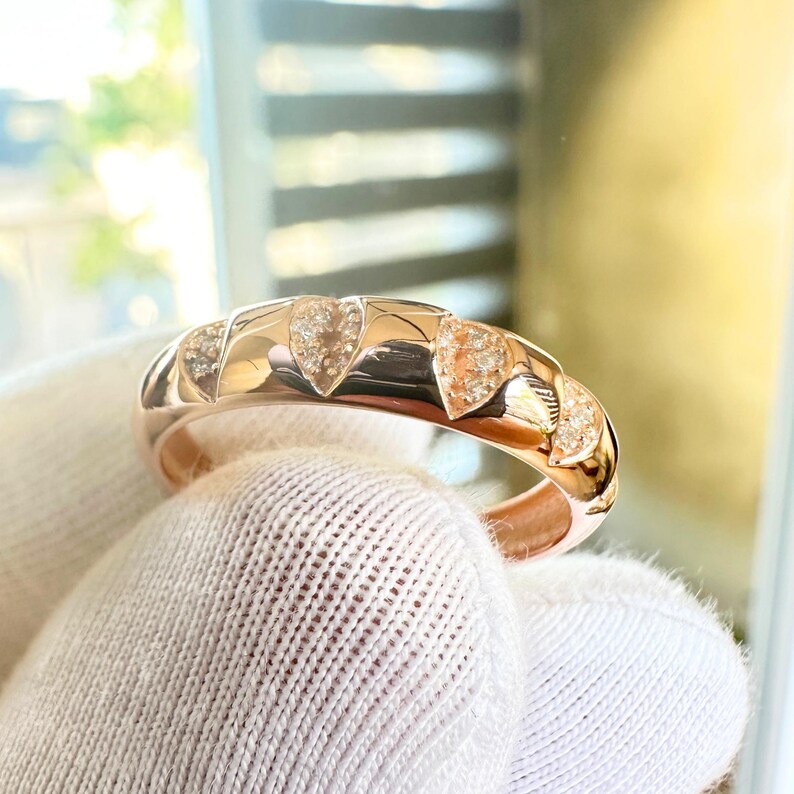 Puede incluir: Anillo de oro rosa con una serie de dise&ntilde;os en forma de coraz&oacute;n, cada uno adornado con peque&ntilde;as piedras brillantes. La superficie pulida del anillo refleja la luz, resaltando los intrincados detalles. El anillo es una joya.