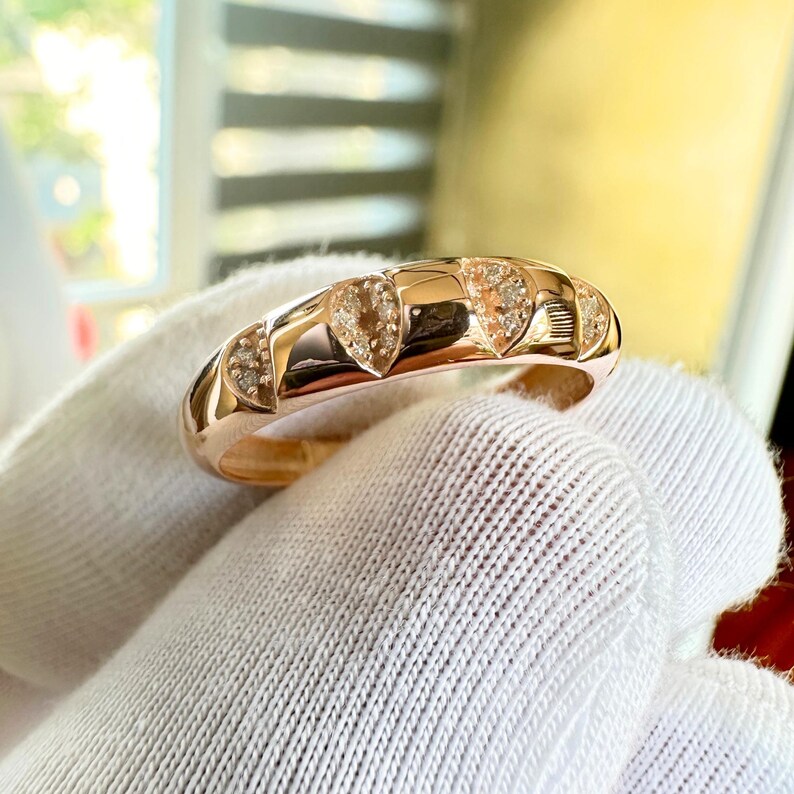 Puede incluir: Un anillo de oro rosa con una serie de dise&ntilde;os en forma de coraz&oacute;n, cada uno adornado con peque&ntilde;as piedras brillantes. El anillo se muestra sobre una superficie blanca, con luz natural que resalta su acabado pulido.