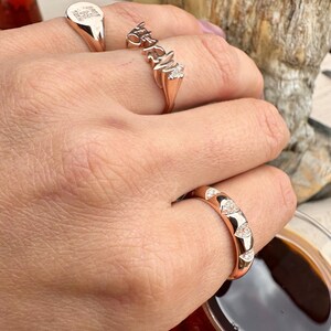 Puede incluir: Una mano que lleva cuatro anillos de oro rosa. Un anillo tiene una cara rectangular con un dise&ntilde;o grabado. Otro anillo deletrea una palabra con peque&ntilde;os diamantes. El tercer anillo tiene un dise&ntilde;o curvo con detalles de diamantes. El cuarto anillo es una banda simple.