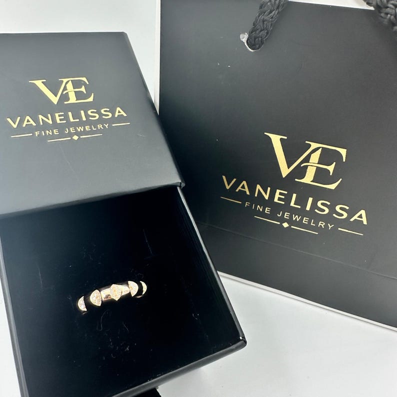 Puede incluir: Un anillo dorado con formas geom&eacute;tricas se muestra dentro de una caja de joyer&iacute;a negra. La caja y una bolsa de compras a juego presentan el texto dorado "VE VANELISSA FINE JEWELRY". El anillo tiene un dise&ntilde;o &uacute;nico con m&uacute;ltiples segmentos.