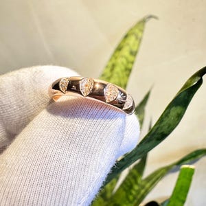 Puede incluir: Anillo de oro rosa con una serie de piedras preciosas claras en forma de pera. El anillo est&aacute; sostenido por una mano enguantada sobre un fondo de follaje verde. Las piedras est&aacute;n espaciadas uniformemente.