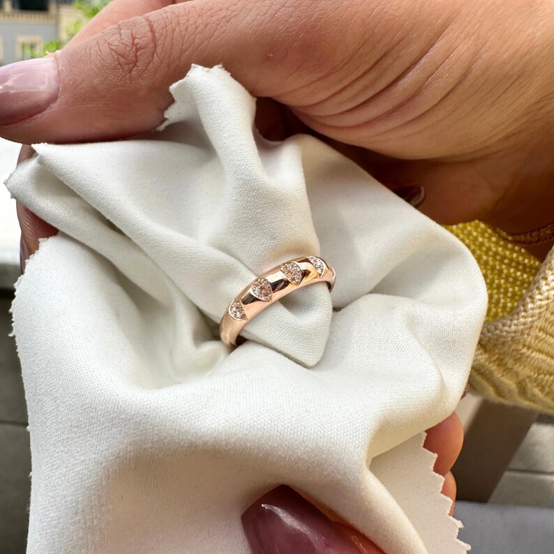 Puede incluir: Anillo de oro rosa con detalles de diamantes en forma de coraz&oacute;n. El anillo se sostiene sobre un pa&ntilde;o blanco. El dise&ntilde;o del anillo presenta una serie de piedras en forma de coraz&oacute;n engastadas a lo largo de la banda, creando un aspecto rom&aacute;ntico y elegante.