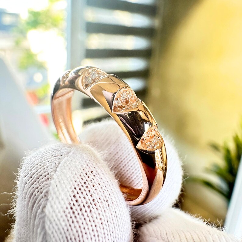 Puede incluir: Anillo de oro rosa con un dise&ntilde;o geom&eacute;trico. El anillo presenta una serie de cortes triangulares, cada uno lleno de peque&ntilde;as piedras brillantes. El anillo es sostenido por una mano enguantada, mostrando su superficie pulida y sus intrincados detalles.