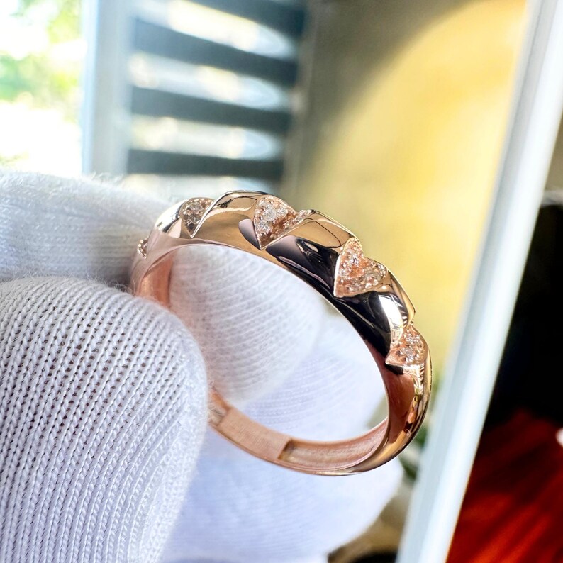 Puede incluir: Anillo de oro rosa con un dise&ntilde;o geom&eacute;trico repetido y peque&ntilde;os detalles brillantes. El anillo tiene un acabado pulido que refleja la luz. El anillo est&aacute; sostenido por una mano enguantada, resaltando su dise&ntilde;o y artesan&iacute;a.