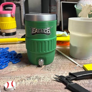 K&ouml;nnte beinhalten: Gr&uuml;ner und silberner Wasserk&uuml;hler mit dem Wort "EAGLES" auf der Vorderseite. Der K&uuml;hler hat einen silbernen Griff und einen Wasserhahn. Im Hintergrund befindet sich ein gelber und rosa K&uuml;hler. Ein baseballf&ouml;rmiges Objekt liegt auf dem Tisch.
