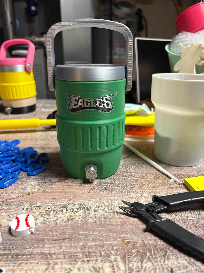 K&ouml;nnte beinhalten: Gr&uuml;ner und silberner Wasserk&uuml;hler mit dem Wort "EAGLES" auf der Vorderseite. Der K&uuml;hler hat einen silbernen Griff und einen Wasserhahn. Im Hintergrund befindet sich ein gelber und rosa K&uuml;hler. Ein baseballf&ouml;rmiges Objekt liegt auf dem Tisch.