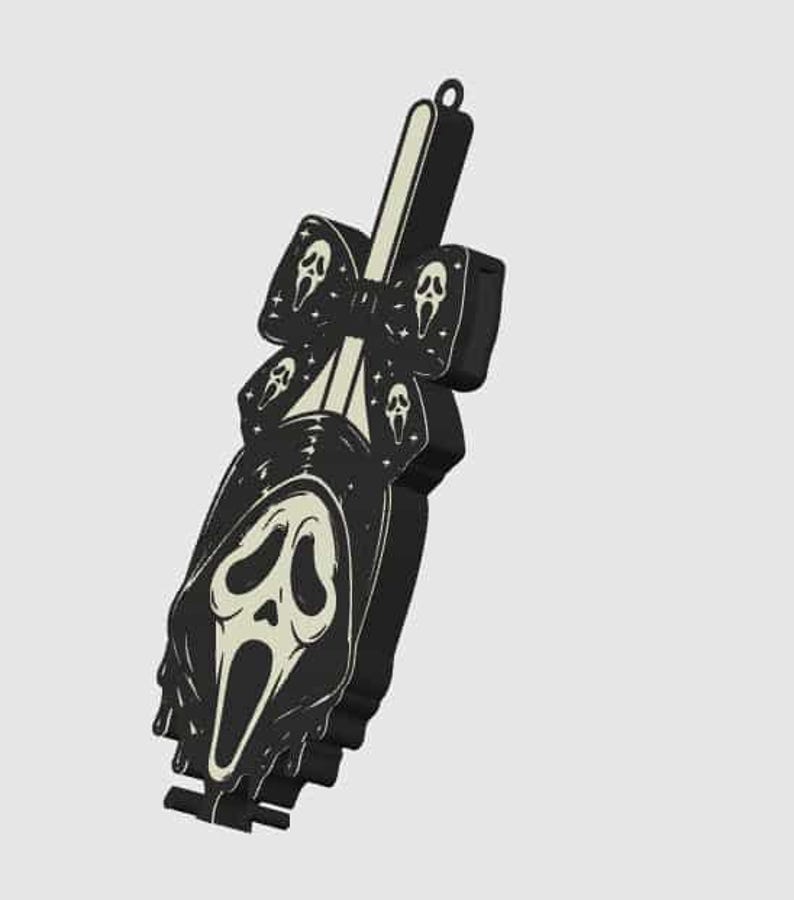May include: A black and off-white decorative accessory featuring a central ghost-like mask graphic with a long, flowing hood. The design includes a bow element adorned with smaller mask icons and tiny star shapes, finished with a small loop at the top.